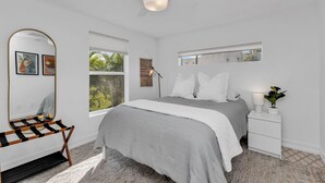 Un dormitorio moderno con una cama, un ventilador de techo, un ventanal grande, un espejo y una mesita de noche con una lámpara.