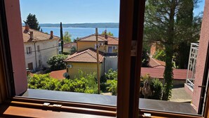 Blick aus einem Fenster auf eine Küstenstadt mit Häusern mit Terrakotta-Dächern und einem tiefblauen Meer im Hintergrund.