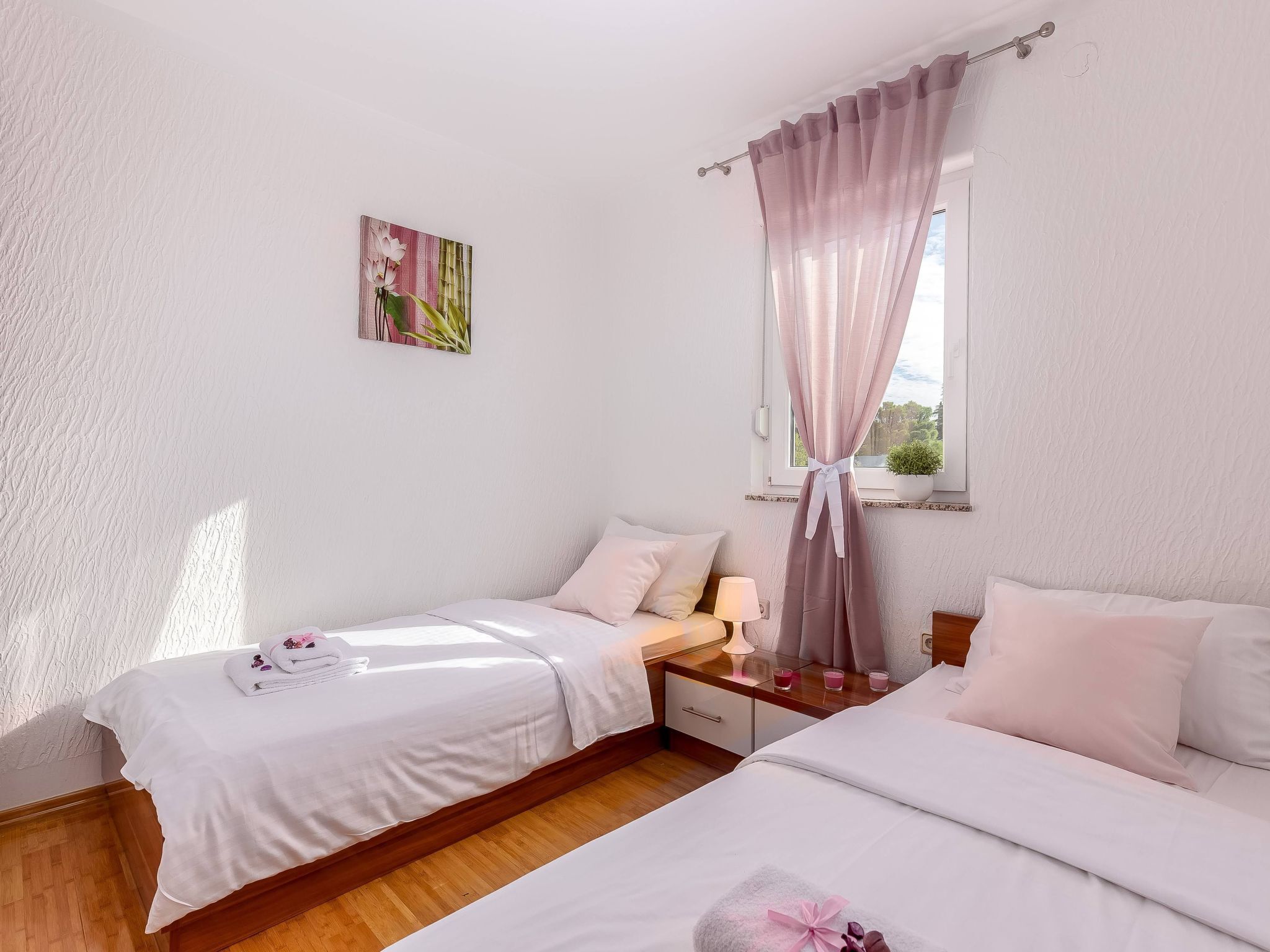A bedroom with two beds, a bedside table with a lamp, a window with curtains, and a wall-mounted picture.