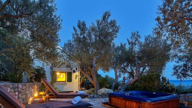 Un espacio al aire libre con suelo de piedra, que cuenta con un jacuzzi, sillones de descanso y un pequeño edificio iluminado, rodeado de árboles y con vistas a un cuerpo de agua.