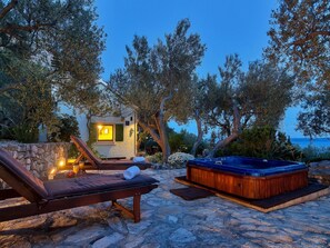 Un espace extérieur dallé de pierres, doté d’un jacuzzi, de chaises longues et d’un petit bâtiment illuminé, le tout entouré d’arbres et offrant une vue sur une étendue d’eau.