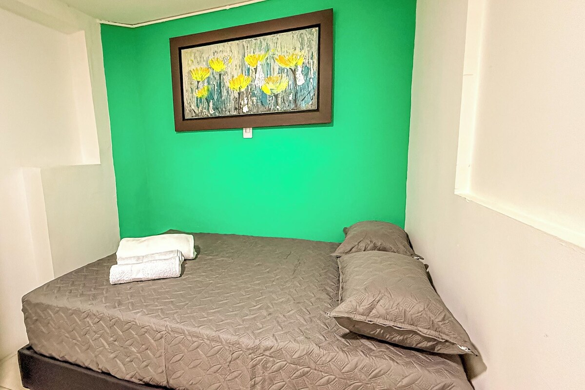 A small, single-bed room with a green wall, a framed painting, and a window.