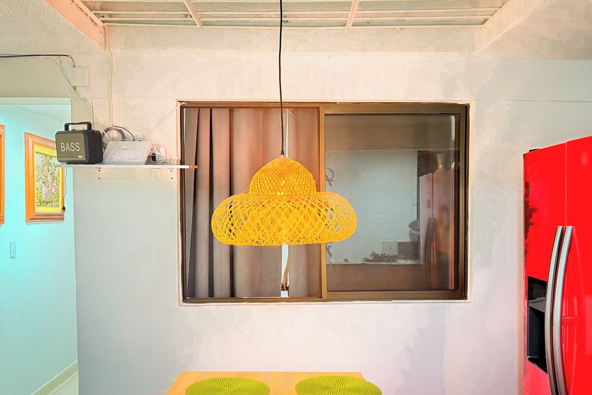 A room with a window, a red refrigerator, and a yellow hanging light fixture.
