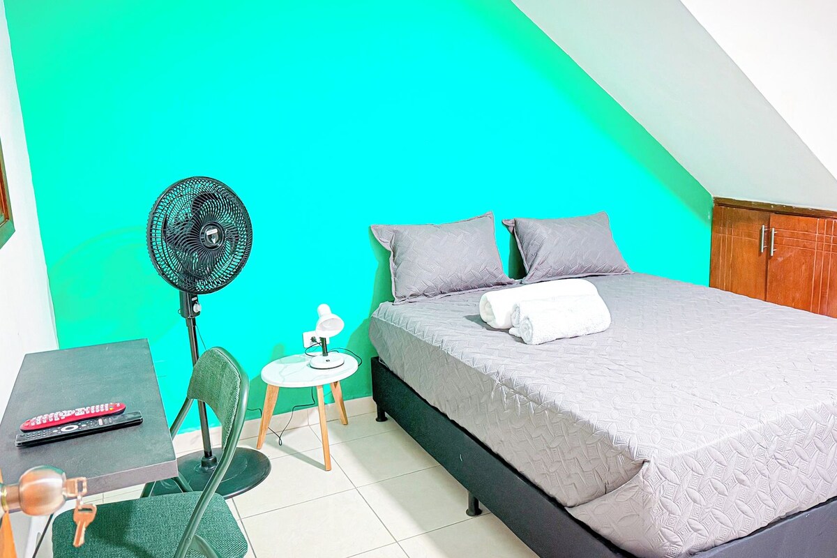 A bedroom with a bed, a desk, a chair, a fan, and a small table.