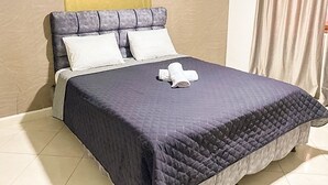 Un lit bien fait, recouvert d’une couverture sombre et agrémenté de deux oreillers blancs, une serviette pliée posée sur le lit, et un mur sobre avec un rideau sur le côté droit.