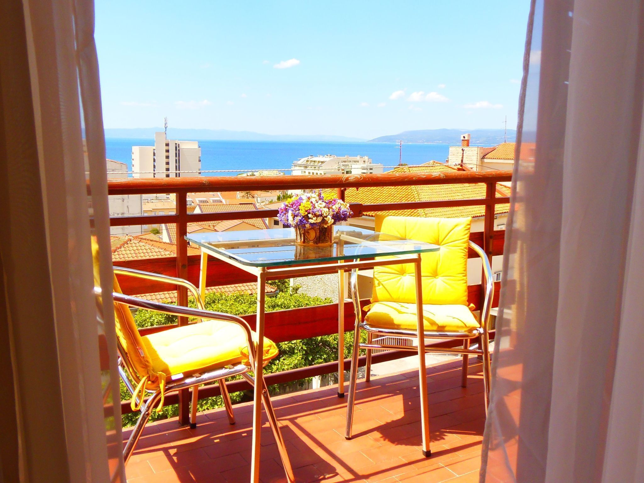 A balcony with a glass table, yellow chairs, and a vase of flowers.