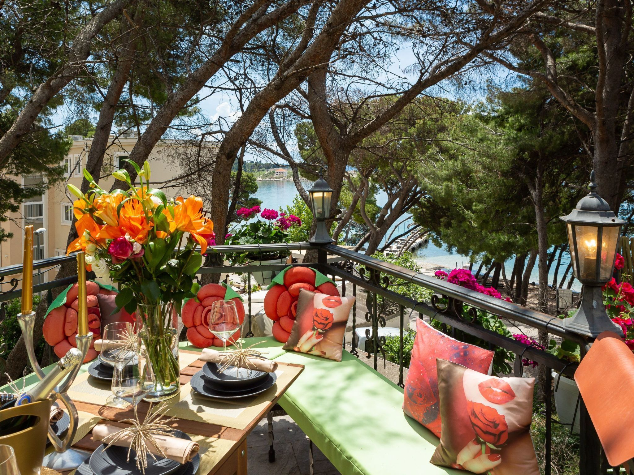 An outdoor dining area with a view of a lake, featuring a table set with plates, glasses, and a vase of flowers.