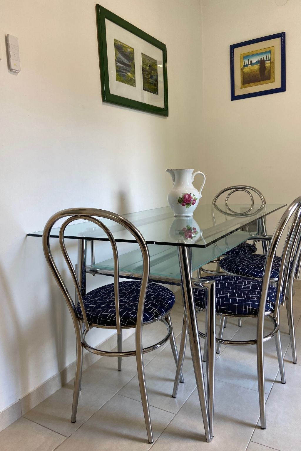 Zona pranzo con tavolo in vetro, sedie in metallo e un decanter bianco decorato con motivi floreali.