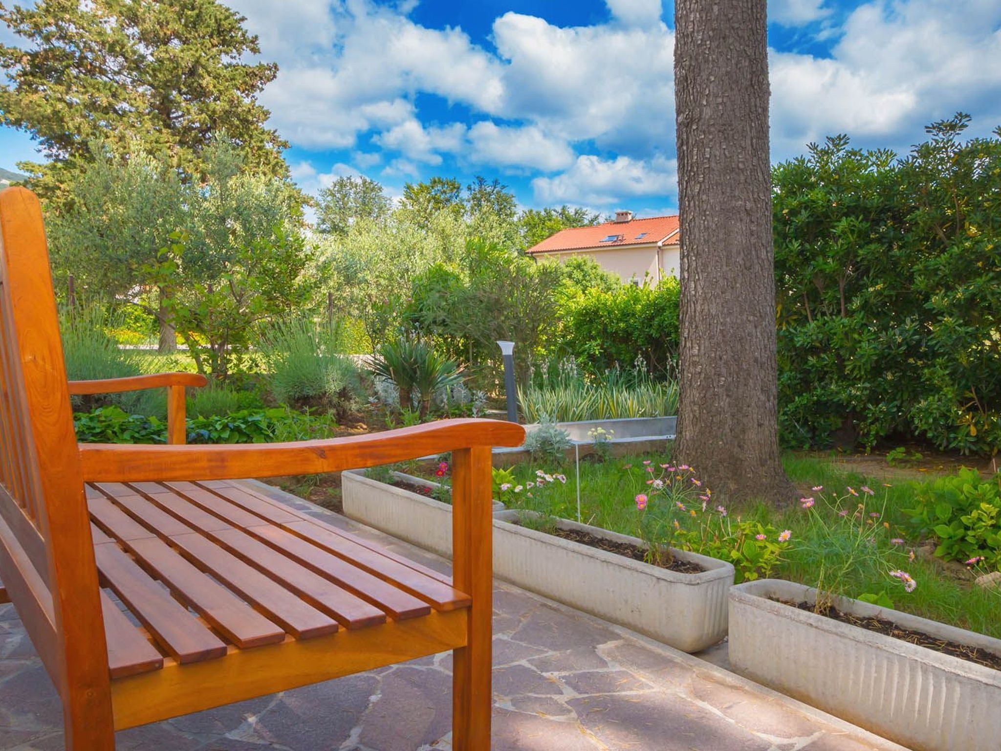 Un banc en bois dans un jardin, avec un sol en pierre, entouré d’une végétation luxuriante et sous un ciel dégagé.