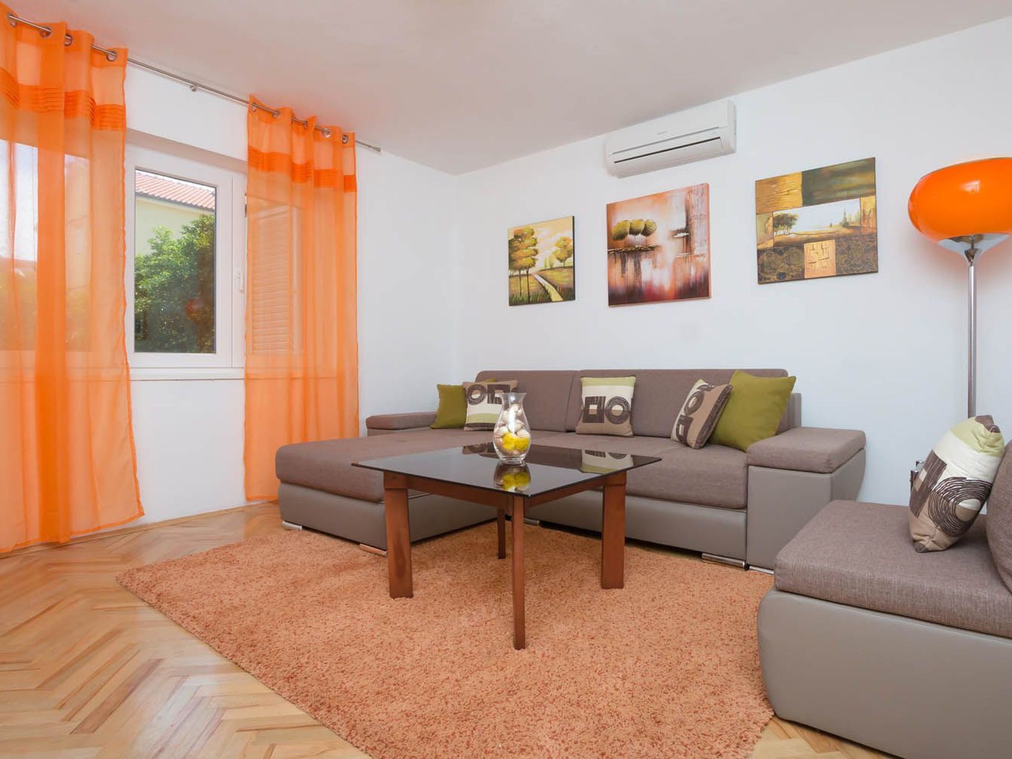 A living room with a sofa, a coffee table, and a rug. There are pictures on the wall and an air conditioner above the sofa.