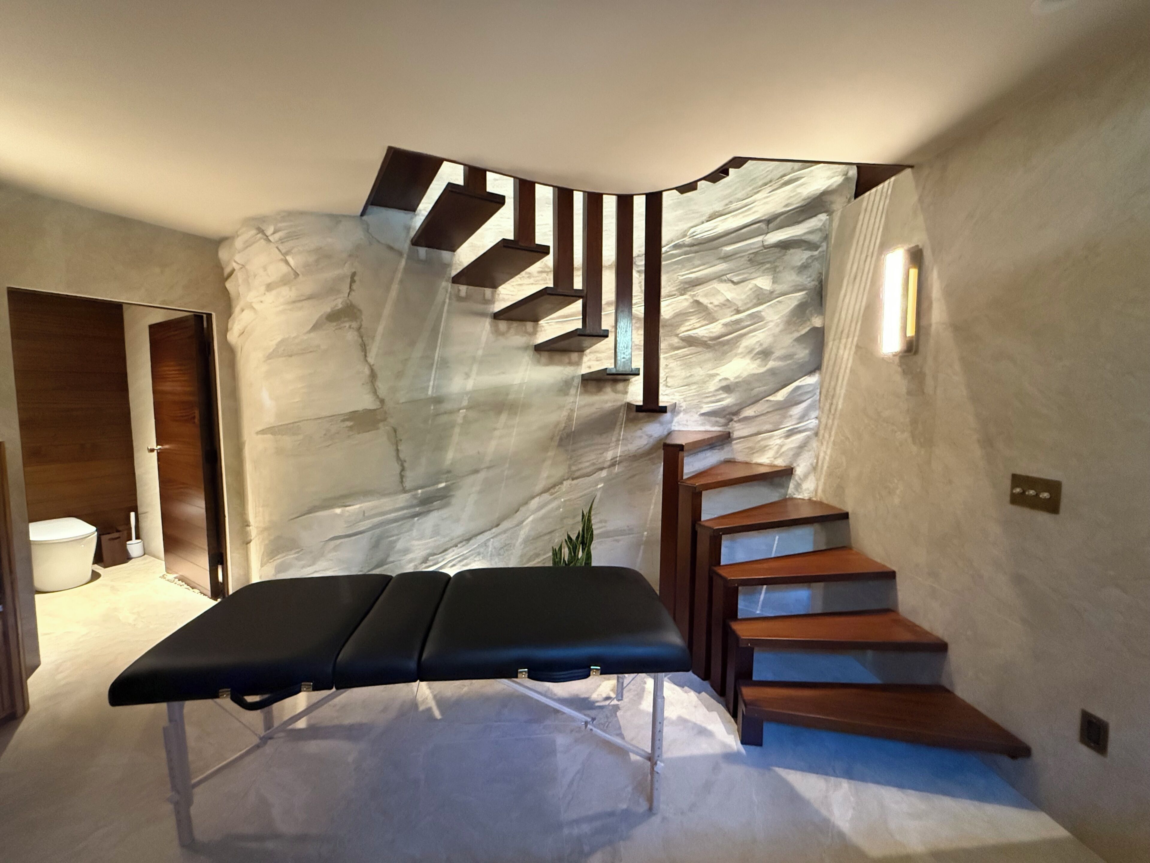 Un interior moderno con una escalera única frente a una pared texturizada, un banco y una puerta que da a un baño.