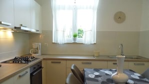 Cocina compacta con estufa a gas, horno y un lavamanos pequeño. Hay una ventana con cortinas translúcidas, un reloj en la pared y una mesa con mantel estampado con hojas.