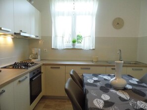 A compact kitchen with a gas stove, oven, and a small sink. There is a window with sheer curtains, a clock on the wall, and a table with a leaf-patterned tablecloth. - Wohnung "Nada" mit Terrasse by Interhome (Krk/Milohnići)