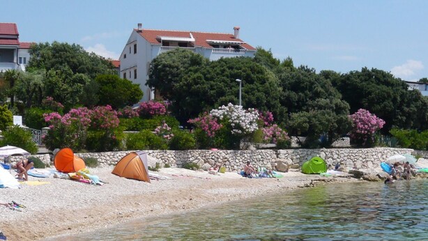 Ein Strand mit Zelten und Menschen, die am Wasser entspannen, einer Steinmauer und einem Haus im Hintergrund.