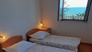 Ein Zimmer mit zwei Betten, einem kleinen Tisch, einem Nachttisch, einem gerahmten Bild an der Wand und einem Fenster mit Meerblick.