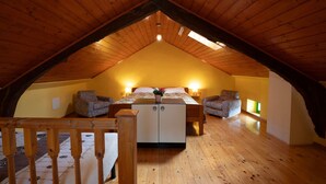 Une chambre mansardée avec un plafond en pente, un plancher en bois, un lit agrémenté d’un arrangement floral, deux fauteuils et une table de chevet avec une lampe.