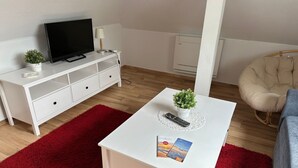 Ein modernes Wohnzimmer mit einem weißen TV-Schrank, einem roten Teppich, einem weißen Couchtisch mit Fernbedienung und Zeitschrift, einem beigen Sessel und einer kleinen Pflanze.