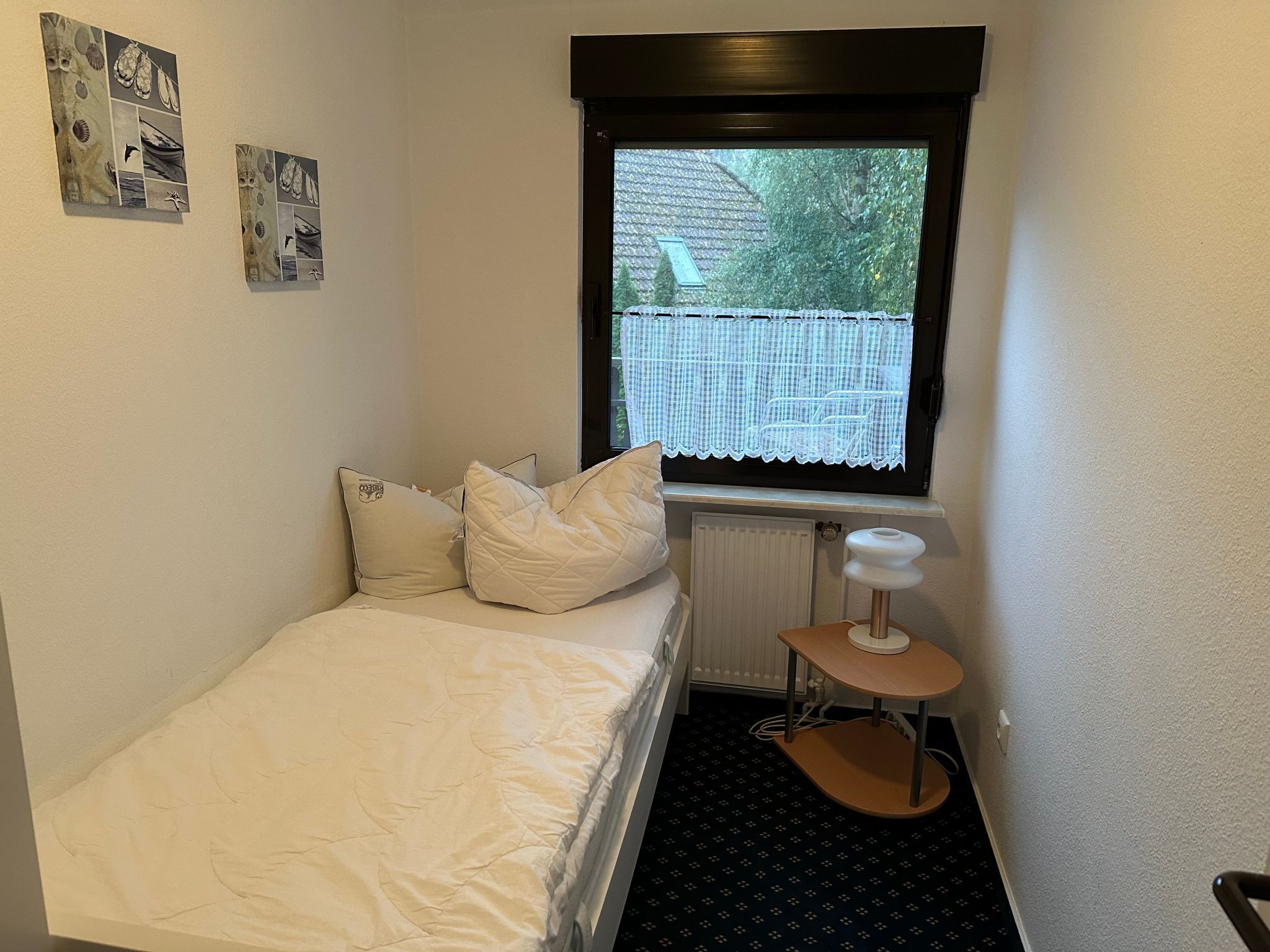 A small, neatly arranged bedroom with a single bed, a window with a view of the outside, and a small table with a chair.