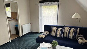 A compact living space with a dark blue sofa, a small white table, a potted plant, and a view of a kitchen through an open door. - Wohnung 1 by Interhome (Ahrenshoop)