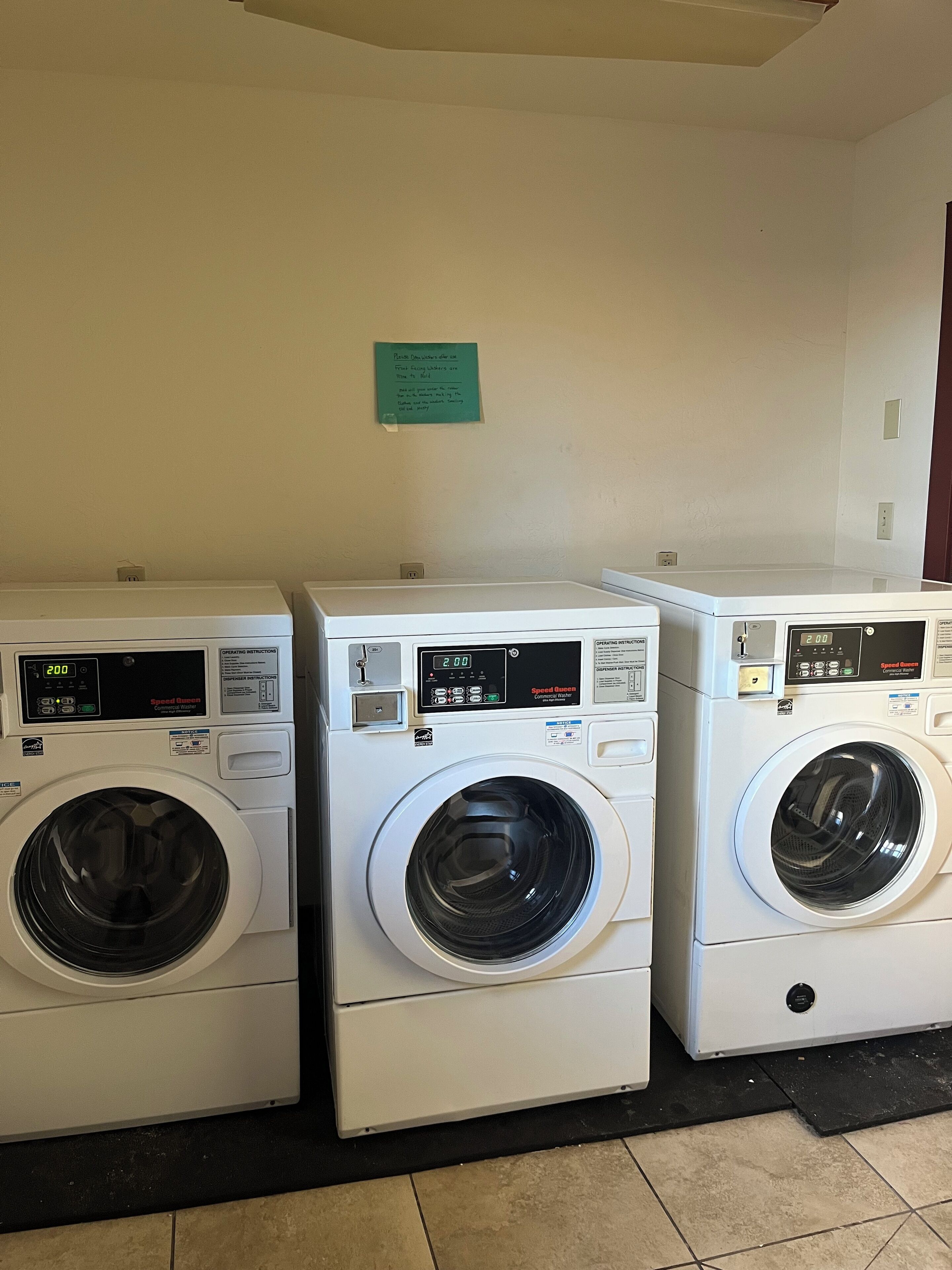 Three stacked front-loading washing machines in a laundry room with a green sign on the wall.