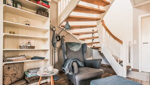 A modern interior with a wooden staircase, a grey armchair, a small round table with a teapot, and a bookshelf filled with various items. - Schwanke´s Feri by Interhome (Gingst)
