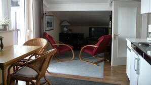Een eetgedeelte met rieten stoelen en een tafel, een keuken met witte kasten, en een rode stoel op de achtergrond.
