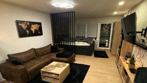 A modern living room with a brown sofa, a wooden coffee table, and a black rug. There is a bed with a white headboard and a window with blinds. A world map is on the wall, and a TV is mounted on the wall. - Winterberg by Interhome (Winterberg)