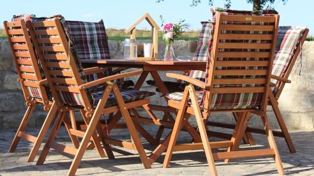 Uma mesa de madeira ao ar livre e quatro cadeiras com almofadas xadrez, dispostas num pátio de calçada com uma parede de pedra e uma árvore ao fundo.