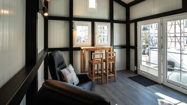 A room with a black leather sofa, a wooden table with chairs, and large windows with blinds. - mit Grill in Bad Marienberg by Interhome (Bad Marienberg)