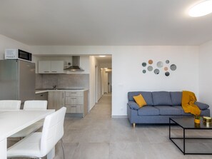 Eine moderne Küche mit integriertem Mikrowellenherd, ein Essbereich mit weißen Stühlen sowie ein Wohnzimmer mit grauem Sofa und Couchtisch.