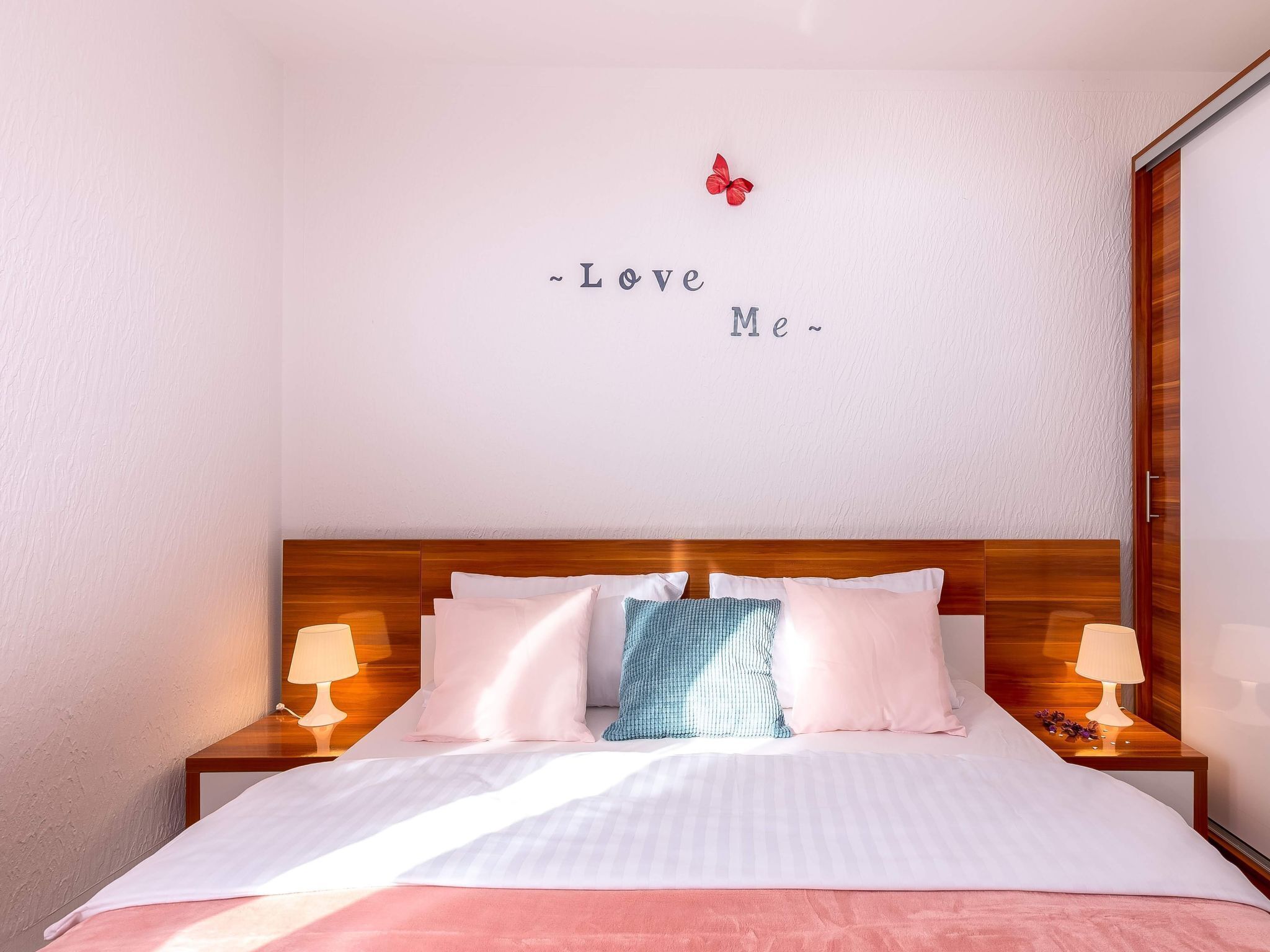 A neatly made bed with a pink blanket, two bedside lamps, and a wall with a decorative butterfly and the words "Love Me" above it.