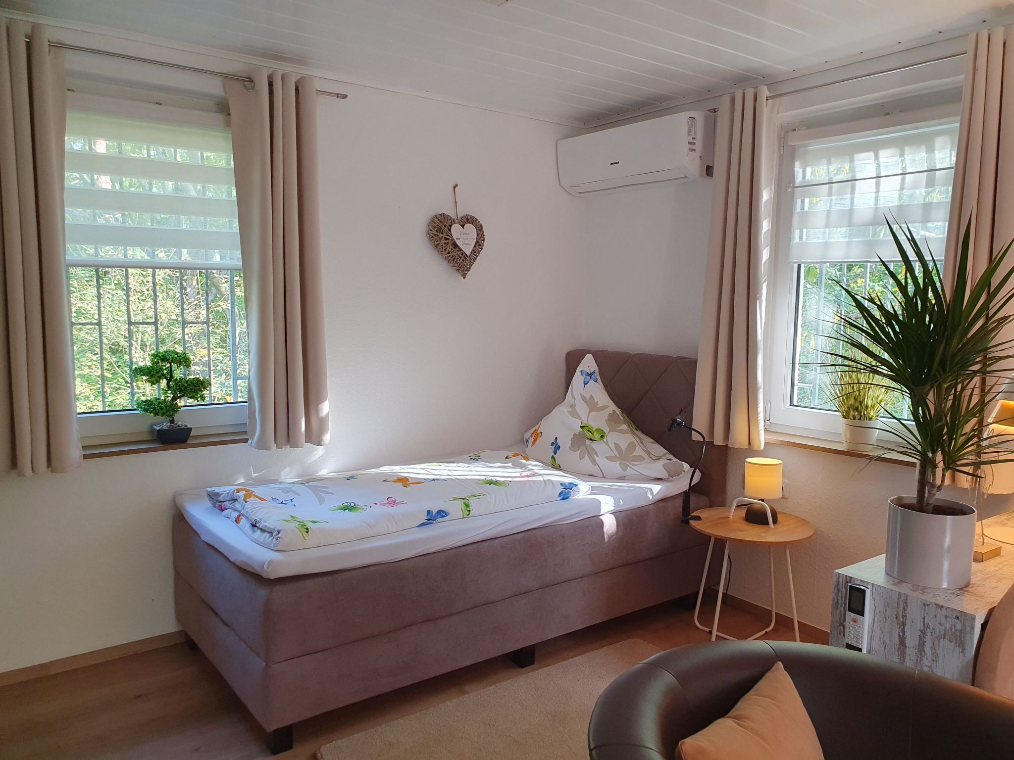 A small, cozy room with a single bed, a bedside table with a lamp, a potted plant, and a wall-mounted air conditioner.