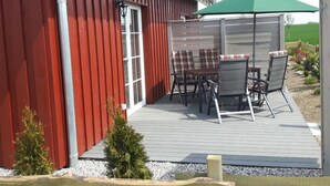 En röd byggnad med en uteplats som har ett bord och stolar, ett grönt parasoll och en liten trädgård.