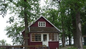 En liten, envåningshus med röd fasad, vit dörr och vita fönster, omgiven av träd och ett trästaket.