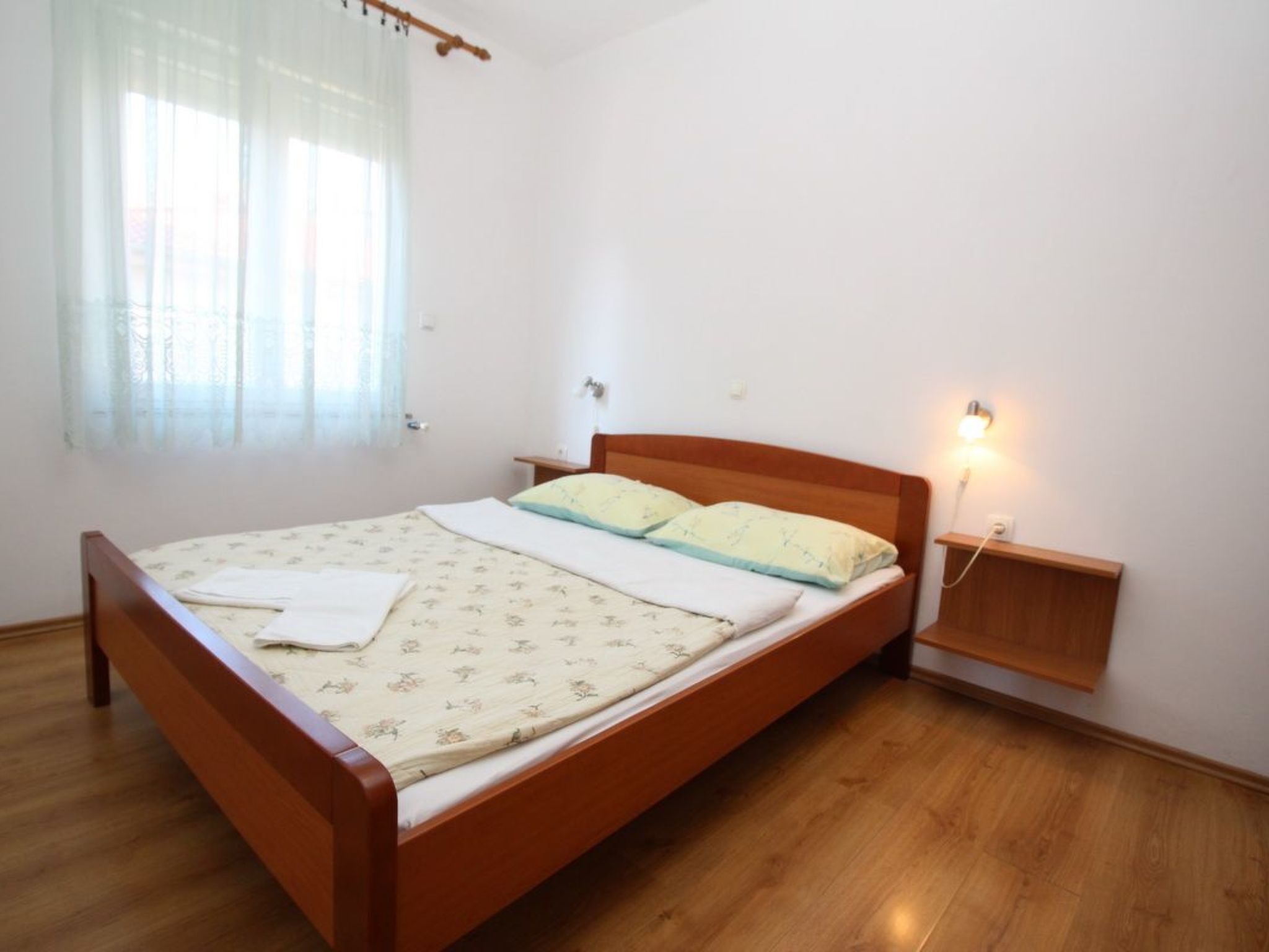 A single bed with a wooden frame, a bedside table, a nightstand with a lamp, and a window with sheer curtains.