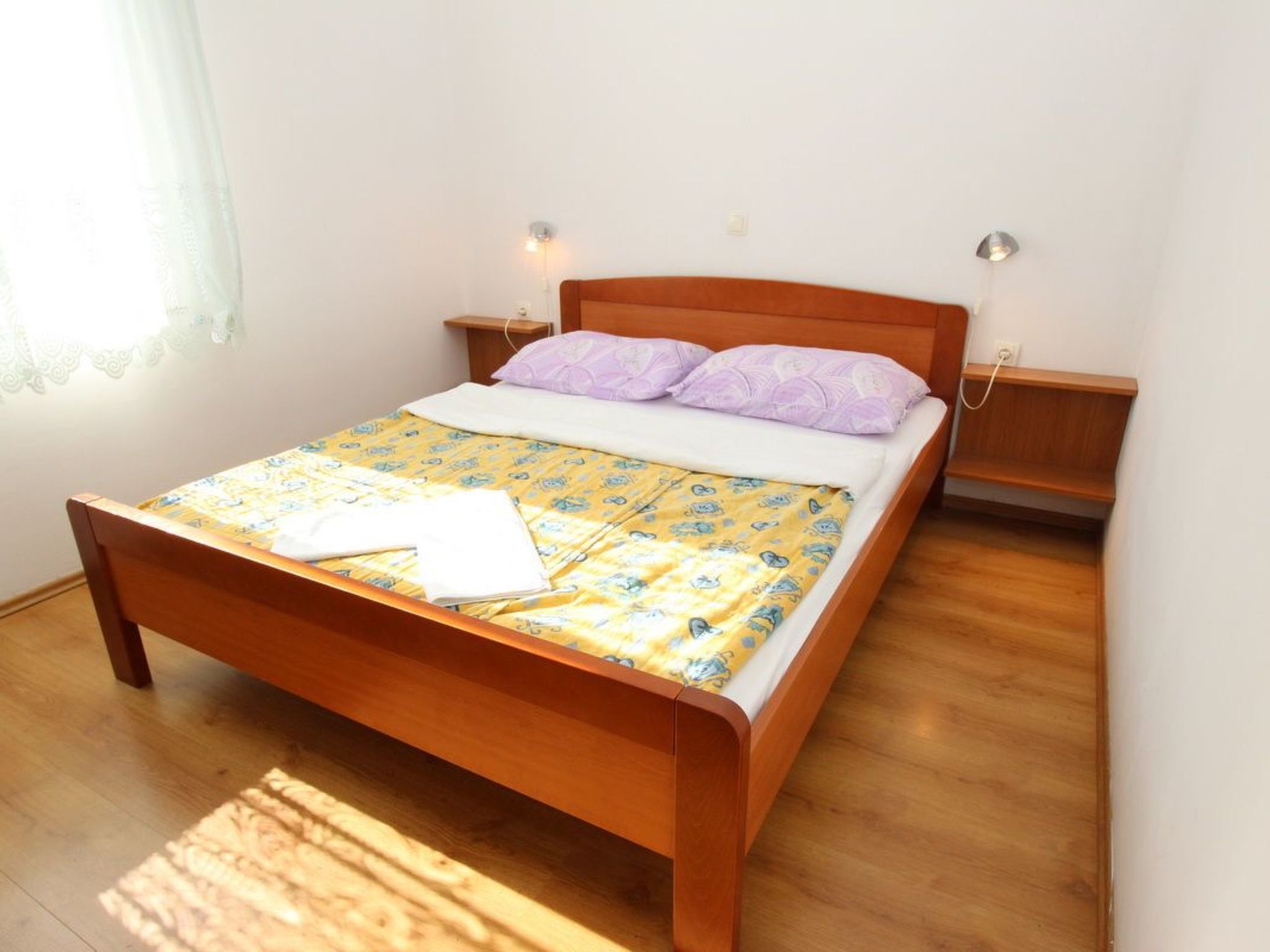 Un lit simple avec un cadre en bois, une couvre-lit à motifs et deux oreillers. Il y a deux tables de chevet avec des lampes, et la pièce est dotée d’un plancher en bois.