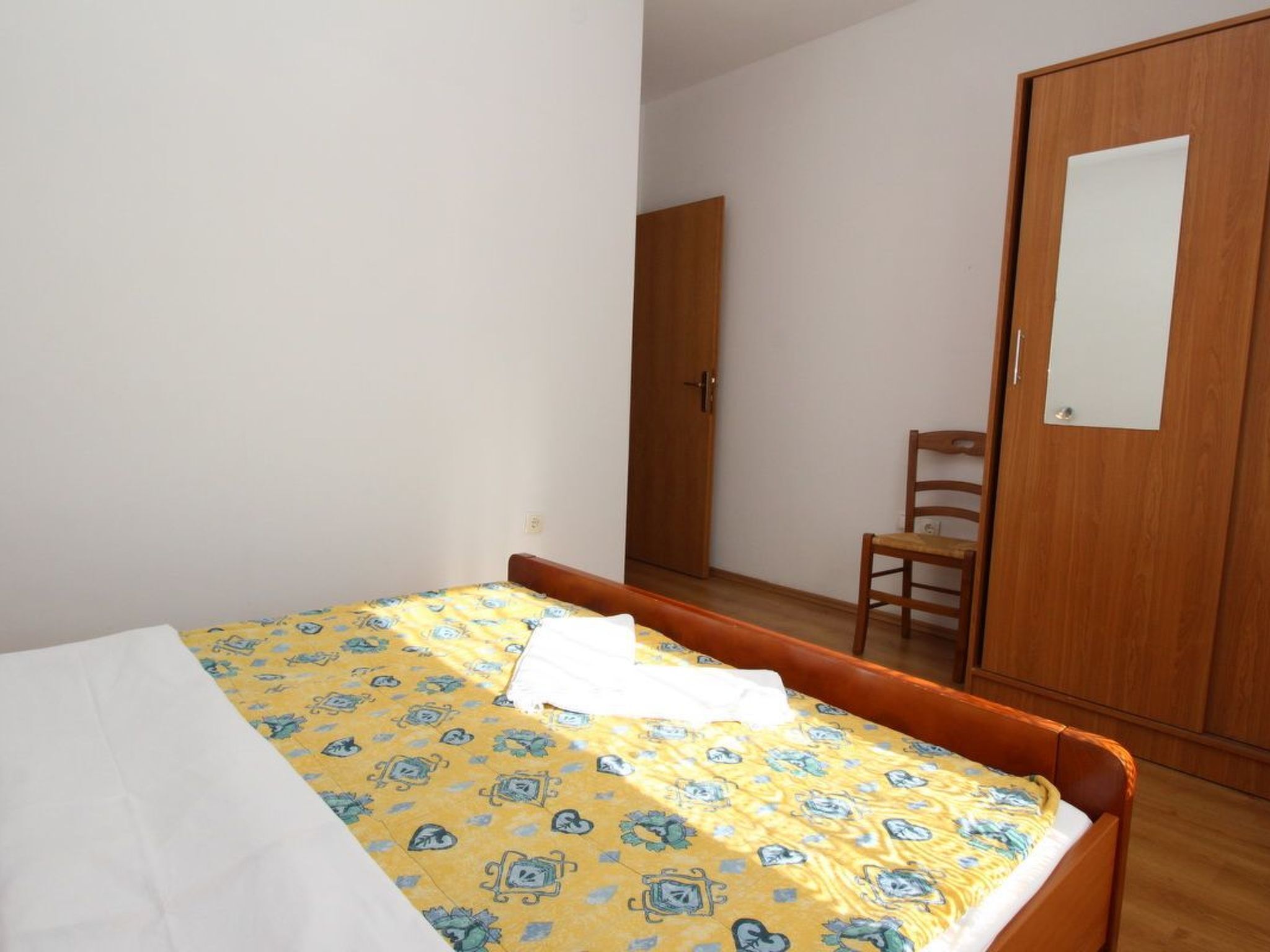 A bedroom with a bed, a wooden wardrobe, a chair, and a mirror.