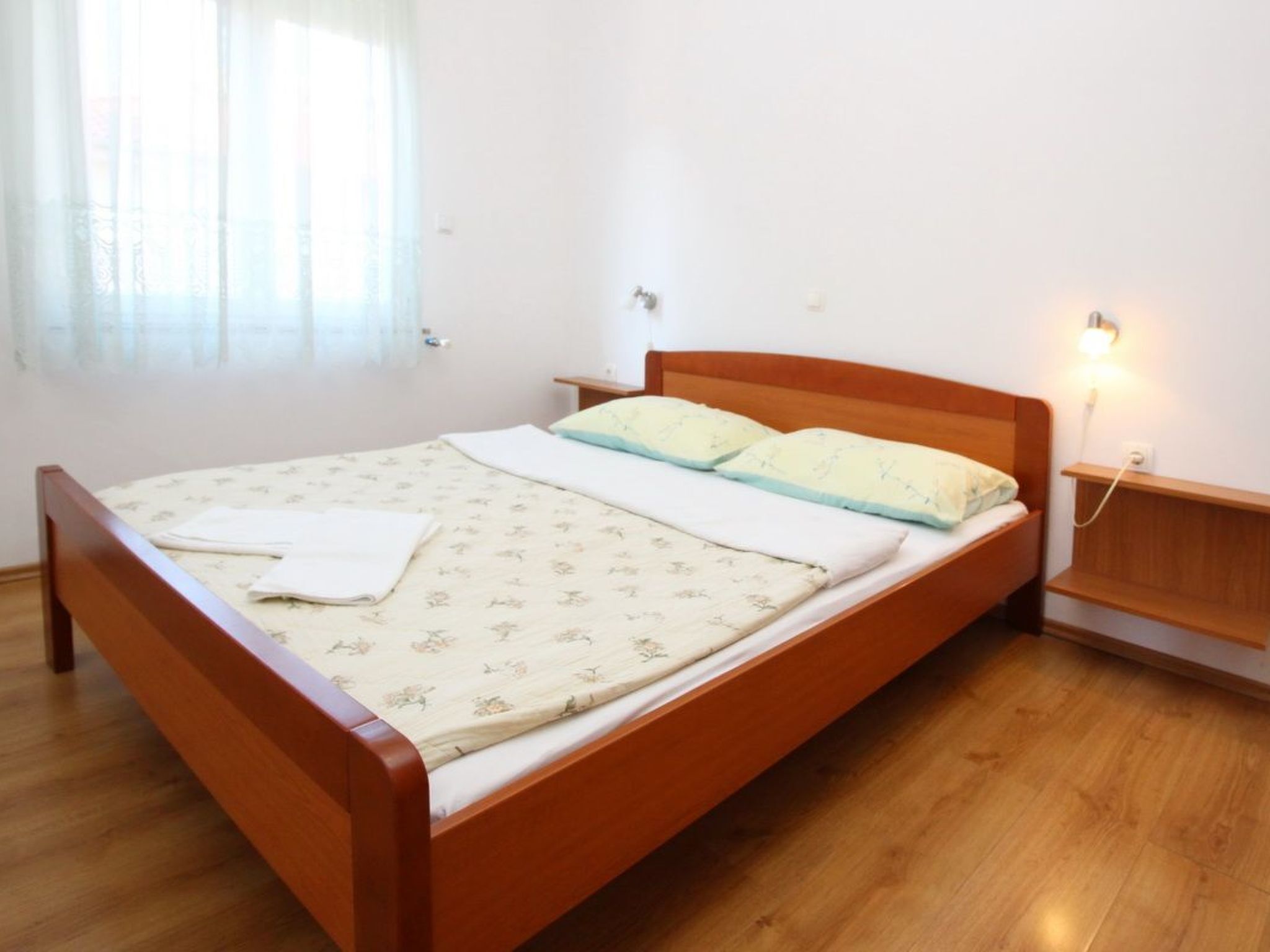 Un letto singolo con struttura in legno, un comodino con una lampada e una finestra con tende leggere.