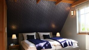 Ein Schlafzimmer mit schrägen Wänden, einem Bett mit weißer Bettwäsche und einer blauen Decke, zwei Nachttischlampen und einem Fenster mit Tageslicht.