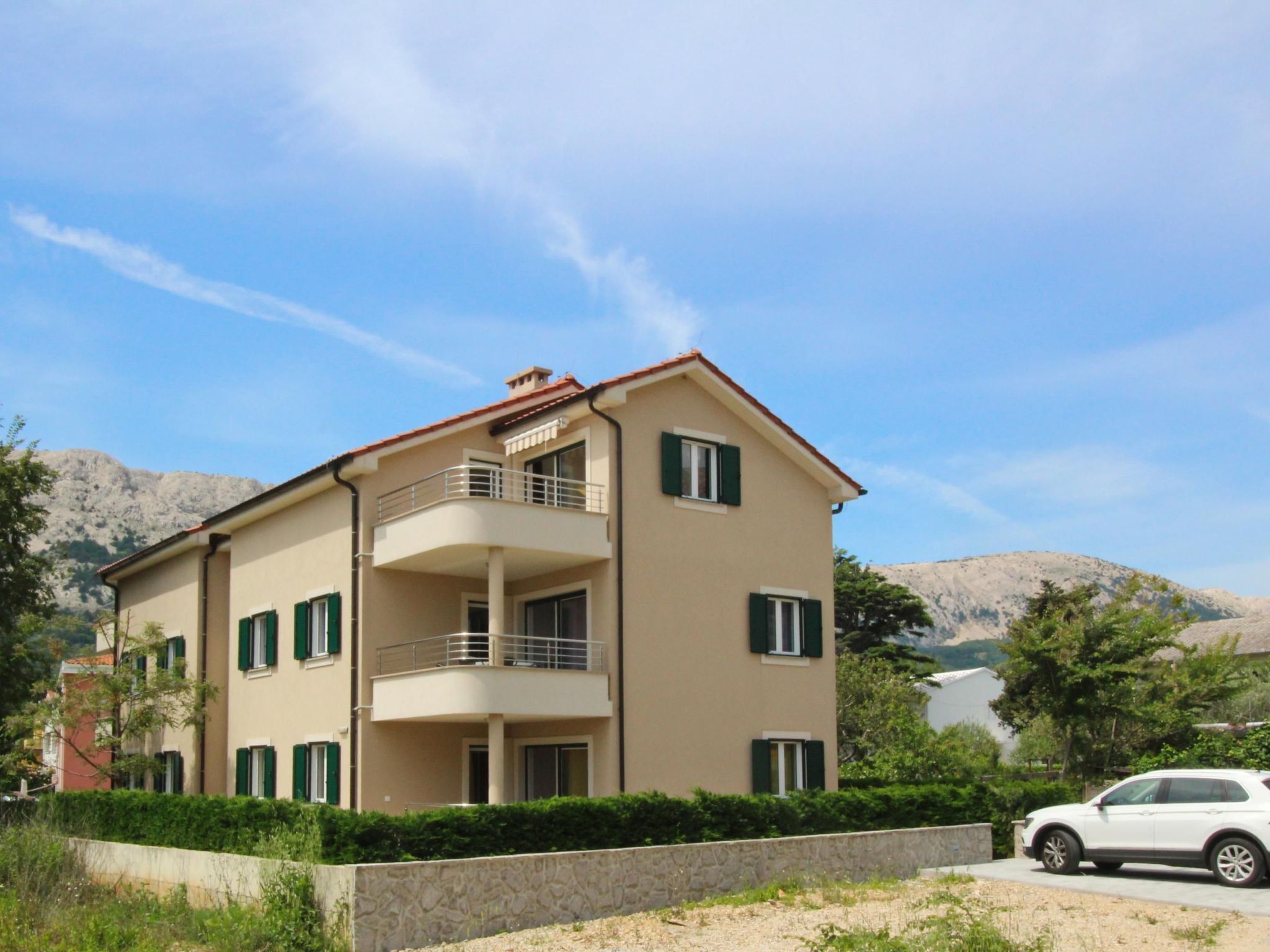A two-story building with a balcony, a stone wall in front, and a car parked nearby.