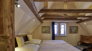 Ein Schlafzimmer mit einem großen Bett, Holzbalken, zwei Fenstern mit Vorhängen und Kunstwerken an den Wänden.