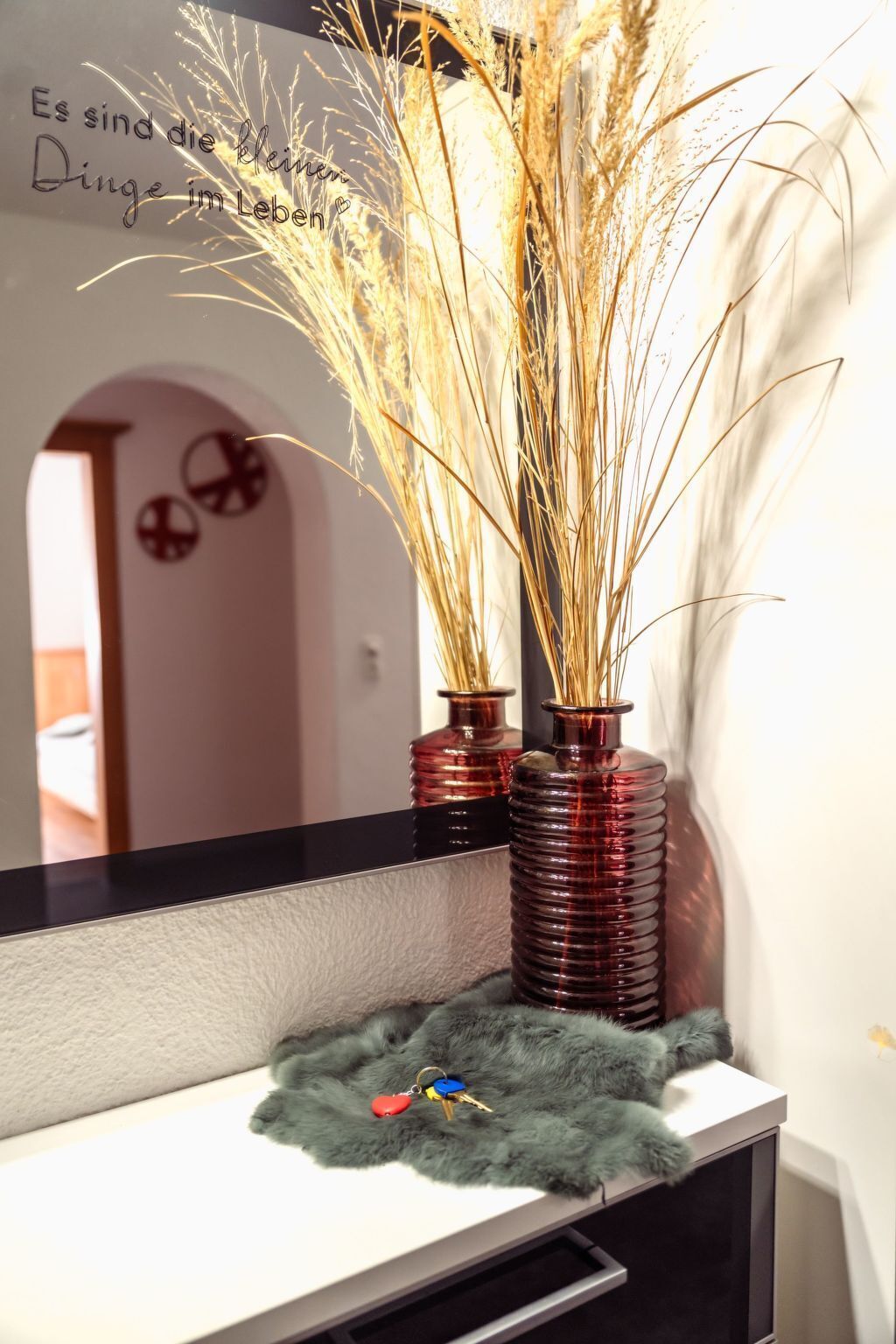 Ein Badezimmer-Waschtisch mit zwei roten Glasvasen, gefüllt mit getrockneten Pflanzen, einem flauschigen grünen Handtuch und einem Spiegel, der eine Tür mit runden Verzierungen reflektiert.