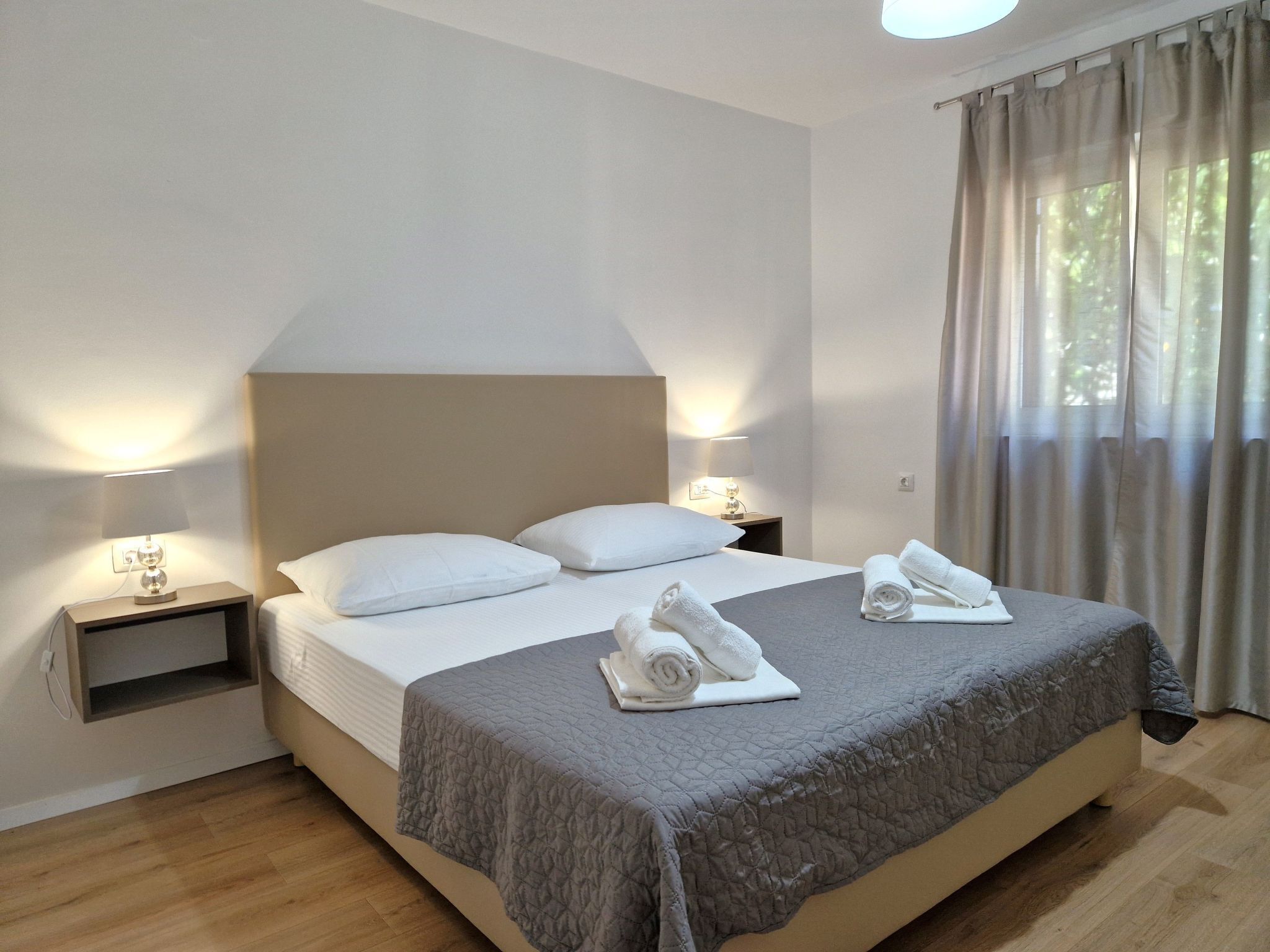 Cama bem arrumada com um edredom cinza, duas almofadas brancas e toalhas dobradas ao lado. Há duas luminárias de cabeceira em mesas de apoio, uma de cada lado da cama. O quarto tem piso de madeira e uma janela grande com cortinas.