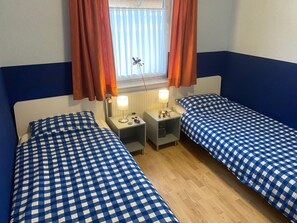 A room with two beds, each with a checkered blue and white bedspread, a window with orange curtains, and a radiator. - Helle Erdgeschosswohnung mit sonniger Terrasse by Interhome (Altfunnixsiel)