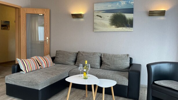 A modern living room with a sectional sofa, a small round table, and a black armchair. There are two wall-mounted lights and a large landscape painting above the sofa. - Helle Erdgeschosswohnung mit sonniger Terrasse by Interhome (Altfunnixsiel)