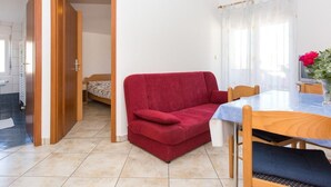 Ein gemütlicher Wohnbereich mit rotem Sofa, einem Holztisch und einer kleinen Küche, die durch eine offene Tür sichtbar ist.