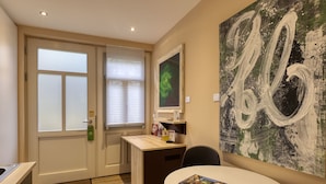 Une chambre d’hôtel compacte comprenant un coin cuisine, un espace repas avec une table et des chaises, un grand tableau abstrait au mur et une porte dotée de panneaux en verre dépoli.