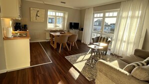 A modern living room with a sofa, a dining table with chairs, a kitchen area, and a large window with curtains. - in Wenningstedt-Braderup (Sylt) by Interhome (Wenningstedt-Braderup)