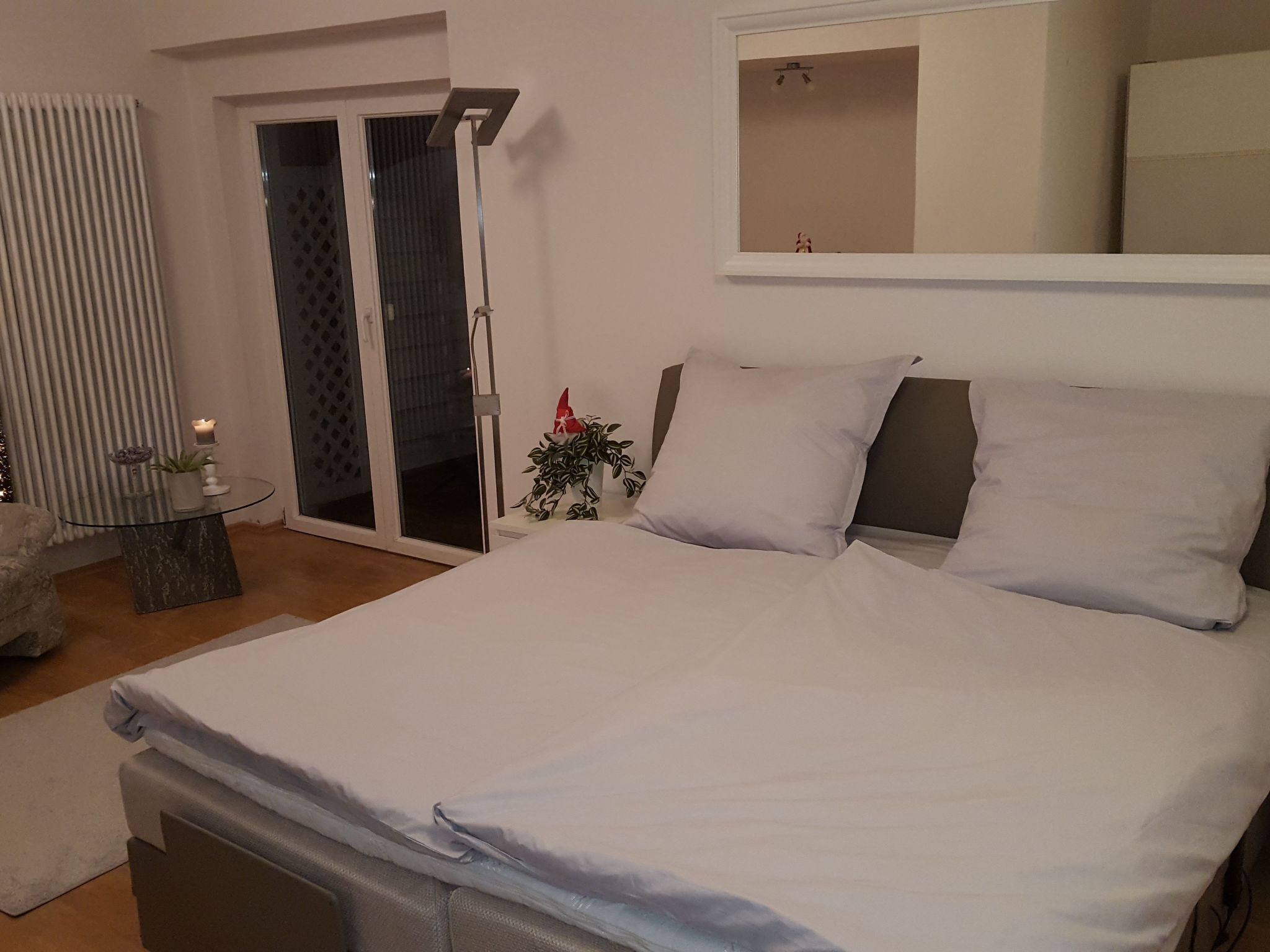 A bedroom with a bed, a small table with a plant, a radiator, and a sliding glass door.