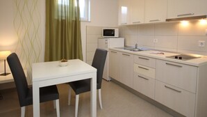 Kompakti keittiö, jossa on valkoiset kaapit, mikroaaltouuni sekä ruokailutila, jossa on pöytä ja tuolit.